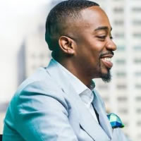 A smiling man in a light blue suit and white shirt sits indoors, facing right, with a blurred skyscraper background.