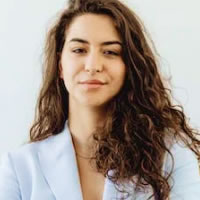 A woman with long, curly hair and a light blue blazer stands against a plain white background, looking straight ahead with a slight smile.