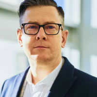 A man with glasses, wearing a suit jacket over a white shirt, looks directly into the camera in a brightly lit indoor space with large windows.