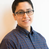 A person wearing glasses and a checkered button-up shirt is smiling while standing against a plain white background.
