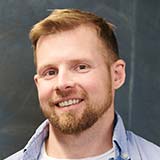 small-testimonial-2 A bearded man with short light brown hair smiles while facing the camera, wearing a light blue collared shirt. The background is a dark, out-of-focus surface.