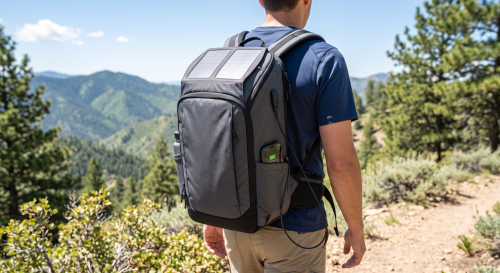 Person wearing a grey backpack with solar panels on a mountain trail, surrounded by evergreen trees and shrubs, overlooking a forested valley below.